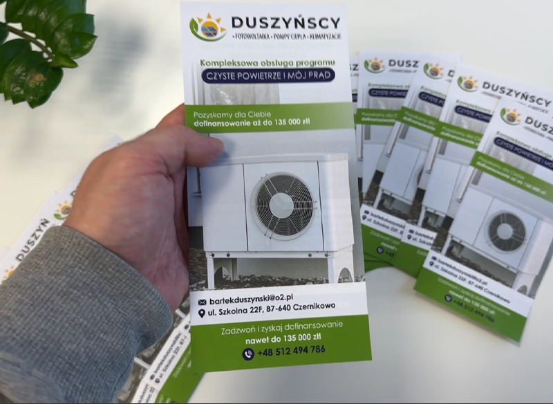 Ulotka reklamowa trzymana w ręce, przedstawiająca jednostkę zewnętrzną pompy ciepła. Ulotki dofinansowanie.