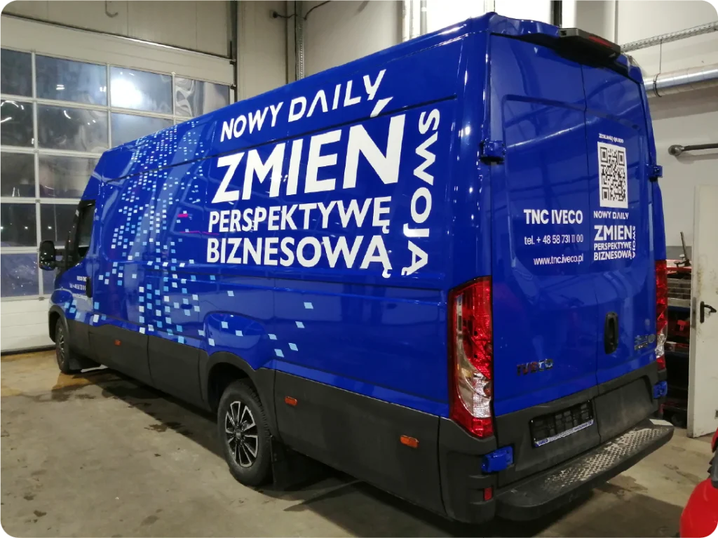 Na załączonym zdjęciu widoczny jest niebieski bus z grafiką reklamową. Na boku i tyle pojazdu widnieje duży, biały napis "NOWY DAILY ZMIEŃ SWOJĄ PERSPEKTYWĘ BIZNESOWĄ". Na tylnych drzwiach po prawej stronie znajduje się logo "TNC IVECO" oraz numer telefonu +48 502 788 100, a także adres strony internetowej www.tnc.iveco.pl. Z prawej strony widoczny jest również kod QR. Na lewym boku busa, w dolnej części, znajduje się grafika składająca się z małych, białych kwadratów, tworzących efekt rozproszenia. Bus stoi w zamkniętym pomieszczeniu lub garażu, na posadzce.