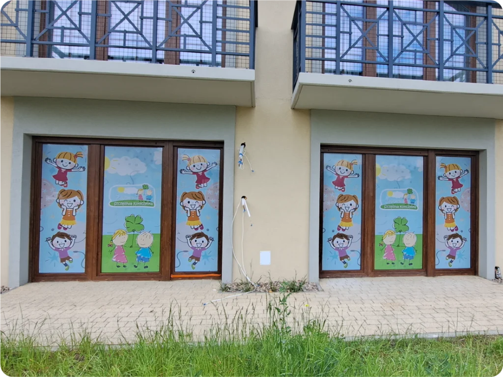 Oklejanie szyb przedszkola folią OWV. Kolorowa grafika reklamowa z postaciami dzieci na witrynie.