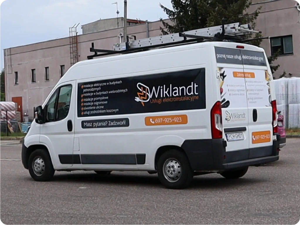 Na załączonym zdjęciu widoczny jest biały bus z reklamą firmy "Wiklandt Usługi elektroinstalacyjne". Na boku pojazdu znajduje się duży, czarny panel z białym logo firmy oraz listą oferowanych usług elektrycznych, takich jak: projektowanie, montaż, modernizacje i pomiary instalacji elektrycznych, instalacje odgromowe, inteligentne domy, monitoring. Poniżej widnieje napis "Masz pytania? Zadzwoń!" oraz pomarańczowy prostokąt z numerem telefonu: 697-925-923. Na tylnych drzwiach busa powtórzono logo i informacje kontaktowe. Na dachu pojazdu zamontowana jest drabina. Bus stoi na utwardzonym podłożu, a w tle widać budynki i ogrodzenia.