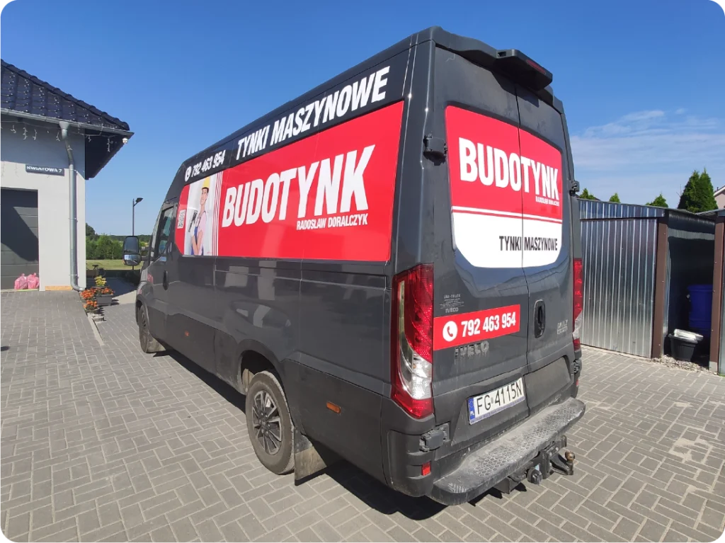 Na załączonym zdjęciu widoczny jest ciemny bus z reklamą firmy "BUDOTYNK" zajmującej się tynkami maszynowymi. Na boku i tylnych drzwiach pojazdu znajduje się duży, czerwony panel z białym napisem "BUDOTYNK" oraz hasłem "TYNKI MASZYNOWE". Obok napisu widnieje zdjęcie mężczyzny, prawdopodobnie właściciela, Dariusza Domalczyka. Na tylnych drzwiach busa powtórzono logo i nazwę firmy, a także podano numer telefonu: 792 463 954. Bus stoi na chodniku wyłożonym kostką brukową, a w tle widać fragment budynku i ogrodzenie.