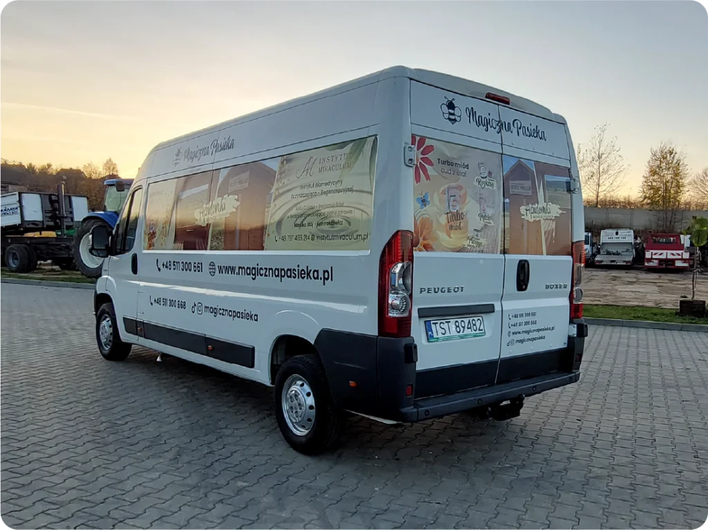 Na załączonym zdjęciu widoczny jest biały bus z grafiką reklamową firmy "Magiczna Pasieka". Na boku i tylnych szybach pojazdu umieszczono naklejki z wizerunkami słoików miodu, plastrów miodu oraz innych produktów pszczelich, a także tekstem informującym o ofercie firmy. Widoczne są również numery telefonów: +48 517 300 563 oraz adres strony internetowej: www.magicznapasieka.pl. Na tylnych drzwiach powtórzono logo firmy. Bus stoi na utwardzonym placu, a w tle widać inne pojazdy i fragmenty budynków.